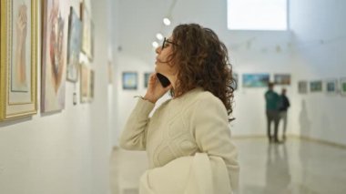 Düşünceli bir kadın, bir sanat galerisini ziyaret ederken telefonla konuşuyor. Serin beyaz bir odadaki tablolara hayran..