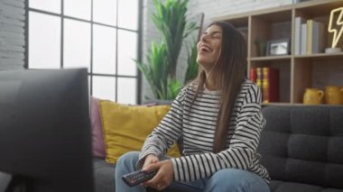 Neşeli bir İspanyol kadın, sıcak bir oturma odasında televizyon seyrediyor, gülüyor ve uzaktan kumandayı tutuyor.