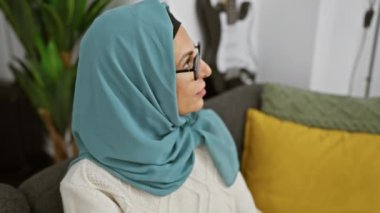 Gülümseyen, orta yaşlı gözlüklü ve tesettürlü bir kadın modern bir oturma odasında rahat bir şekilde oturur, sıcaklık ve tarz yayar..