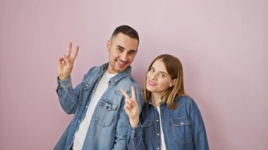Mutlu bir çift, kot gömleklerle kazanan, parmaklarıyla zafer işareti gösteren, izole edilmiş pembe arka planda gülümseyen