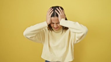 Genç ve güzel İspanyol kadın ayakta duran kazak giyiyor. Baş ağrısı, çaresiz ve stresli. Ağrı ve migren yüzünden. Ellerini başının üstüne koy. izole edilmiş sarı arkaplan