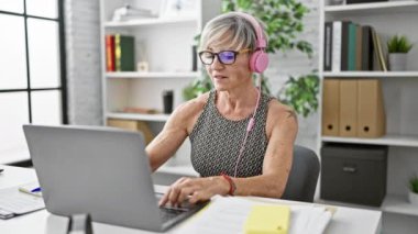 Ofis ortamında kulaklık takıp dizüstü bilgisayar kullanan gri saçlı, olgun İspanyol bir kadın..