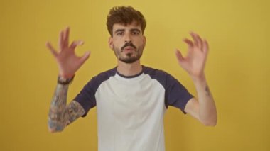 Genç İspanyol bir adam izole edilmiş sarı bir arka plana karşı duruyor, dilini uzatıyor ve parmaklarının arasından bakıyor, neşeli, olumlu bir ifade yaratıyor..