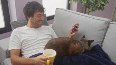 Gülümseyen bir adam, elinde kahve fincanı tutarken bir siyam kedisini okşayıp akıllı telefon kullanıyor..