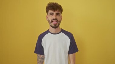 Mutlu bir ifade, genç İspanyol adam izole edilmiş sarı bir arka planda duruyor, komik, eğlenceli bir duygu gösterisiyle dilini dışarı çıkarıyor.