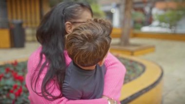 Renkli bir parkta sevgiyle sarılan gülümseyen bir kadın ve çocuk anneliği, aileyi ve neşeyi simgeliyor..