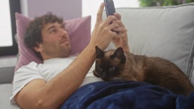 Rahat bir adam rahat evinde siyam kedisiyle kanepede akıllı telefon kullanıyor.