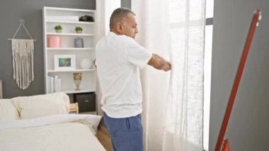 Gündelik giysiler içindeki orta yaşlı bir İspanyol şık dekorlu modern bir yatak odasında perdeleri ayarlıyor..