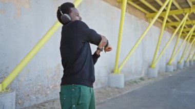 Afro-Amerikalı bir adam şehir ortamında kulaklıklar ve günlük kıyafetlerle esniyor..