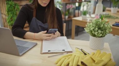 Çiçekçide dizüstü bilgisayarla çalışırken akıllı telefon kullanan odaklanmış bir İspanyol kadın.