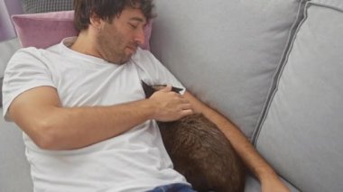 Beyaz gömlekli genç bir İspanyol, gri bir kanepede kahverengi kedisiyle rahat bir evde kestiriyor..