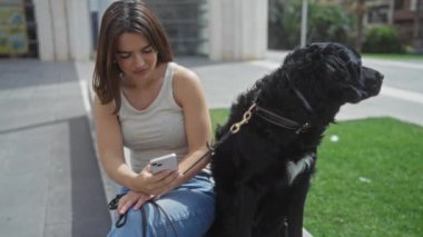 Genç bir İspanyol kadın akıllı telefonunu kullanırken evcil köpeği siyah bir labrador ile şehir parkında oturuyor..