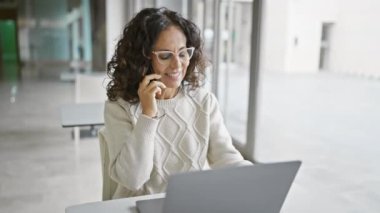 Gözlüklü olgun bir İspanyol kadın modern bir ofiste dizüstü bilgisayar ve akıllı telefon kullanıyor..