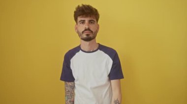 Kendinden emin, genç İspanyol bir adam izole edilmiş sarı bir arka plana karşı duruyor, bir gözünü eliyle kapatıyor, gülümseyen bir yüz takınıyor.