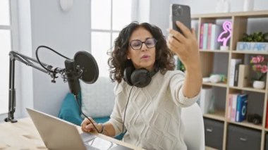 İçerde selfie çekerken mikrofon ve dizüstü bilgisayarla podcast stüdyosunda yaşayan İspanyol kadın..