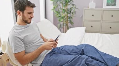 Sakallı, Latin kökenli genç bir adam rahat bir yatak odasında akıllı telefon kullanıyor..