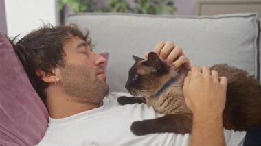 İçerik sahibi İspanyol bir adam evde gevşiyor, şefkatli siyam kedisini rahat bir kanepede okşuyor..