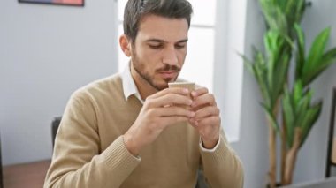 Düşünceli, genç, İspanyol bir adam modern ofis ortamında kahve molası verip üretkenliği ve tefekkürü çağrıştırıyor..
