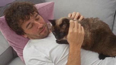 Genç bir adam, rahat bir oturma odasında gri bir kanepede dinlenirken bir siyam kedisini sevgiyle besliyor..