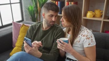 Bir çift, rahat bir oturma odasında birlikte vakit geçirmeyi seviyor. Akıllı telefonlu bir adam, elinde kupa tutan bir kadın..