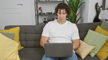 Oturma odasındaki kanepede oturmuş evinde bilgisayarında neşeyle internette sörf yapan sırıtan genç İspanyol bir adam.