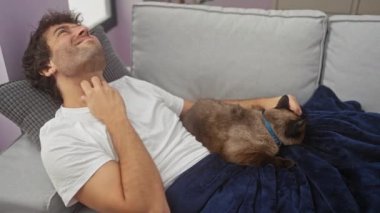 Genç bir İspanyol adam kucağında siyam kedisiyle kanepede yatarken boynunu kaşır, sıcak bir oturma odasında yakalanır..