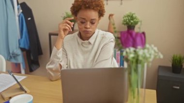 Evinde çalışan siyah bir kadın, dizüstü bilgisayarının önünde stres altında. Evrakları, gözlükleri ve ev bitkileriyle..