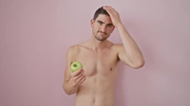 Kendine güveni tam bir İspanyol genç, doğal bir poz veriyor, üstsüz ve elmalı, pembe izole arka planda, rahat, mutlu bir gülümseme yayıyor.