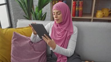 Pembe tesettürlü bir kadın, iyi ışıklandırılmış, rahat bir oturma odasındaki kanepede dinlenirken dikkatle tablete odaklanır..