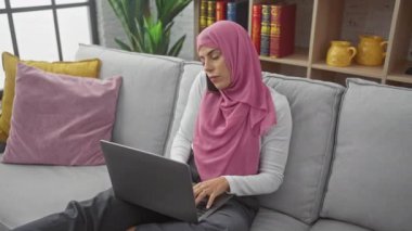 Modern bir evde pembe tesettür, dizüstü bilgisayar ve akıllı telefon takan odaklanmış bir kadın..