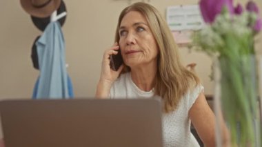 Beyaz kazaklı olgun bir kadın evde cep telefonuyla konuşuyor. Etrafı kitaplarla ve ev bitkileriyle çevrili..