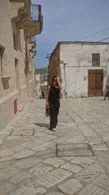 Latin kökenli genç bir kadın eski bir kasabada yürüyor, Mata, Basilicata, İtalya, Avrupa, siyah giyinmiş, örülmüş bir çanta tutuyor ve gülümsüyor tarihi taş döşeli caddeleri keşfederken.