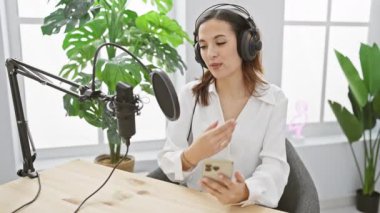 Genç bir kadın, kapalı bitki dolu bir stüdyoda akıllı telefon tutarken mikrofonla bir podcast kaydediyor..