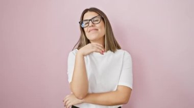 Kendine güvenen genç İspanyol kadın pembe arka planda kollarını kavuşturarak poz veriyor, akıllı ve pozitif, gözlük takıyor ve kameraya gülümsüyor.