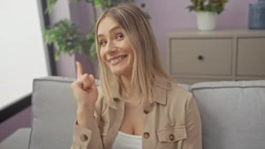 Kanepede oturan genç sarışın kadın mutlu ve komik yüzüyle kamerayı işaret ediyor..