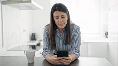 Genç, güzel, İspanyol bir kadın mutfakta tezgaha yaslanmış akıllı telefon kullanıyor.