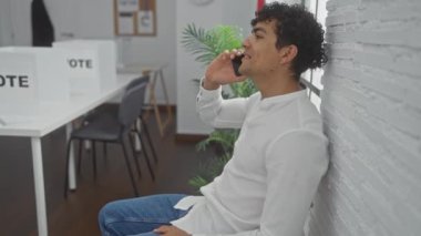 Beyaz tişörtlü İspanyol adam oy pusulası olan parlak bir oy odasında telefon kullanıyor.