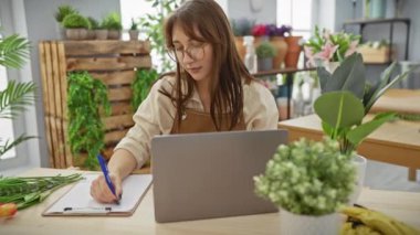 Genç bir kadın dizüstü bilgisayarı, bitkileri ve panosu olan bir çiçekçide not alıyor..