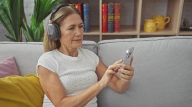 Kafkasyalı olgun bir kadın rahat bir oturma odasında kulaklığı ve akıllı telefonuyla müzik dinliyor..
