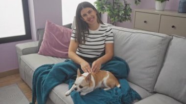 Genç bir İspanyol kadın sıcak bir oturma odasında Chihuahua köpeğiyle oturuyor, gülümsüyor ve elleriyle kalp şekli yapıyor..