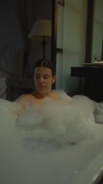 Huzurlu genç bir kadın loş bir otel odasında köpük banyosunun keyfini çıkarıyor. Rahatlık ve lüksün vücut bulmuş hali..