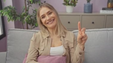 Kanepede oturmuş gülümseyen ve kameraya göz kırpan genç sarışın kadın parmaklarıyla zafer işareti yapıyor. İki numara. evde.