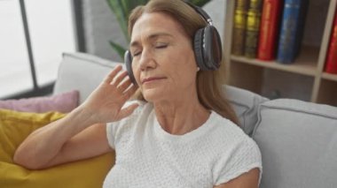 Kulaklık takan olgun bir kadın rahat bir oturma odasındaki kanepede müzik dinlemekten hoşlanır..
