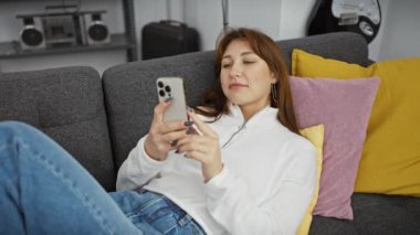 Kafkasyalı rahat bir kadın modern oturma odasındaki koltukta akıllı telefon kullanıyor.