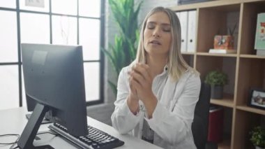 Stresli görünen genç bir kadın doktor, bir klinikte ya da hastanenin iş yerinde masa başında oturur ve kapalı alandaki bilgisayar ekranına odaklanır..