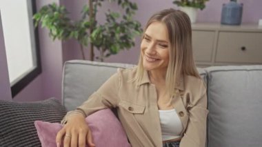 Kanepede oturan genç sarışın kadın selam veriyor mutlu ve gülümsüyor, evine hoş geldin jesti yapıyor.