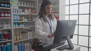 Genç bir kadın eczacı tamamen dolu bir eczanenin tezgahında çalışırken bilgisayar kullanıyor..