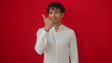Tişört giyen, ayakta duran ve gülümseyen mutlu, genç İspanyol bir adam. Ayrı bir kırmızı arka plan üzerinde başparmağıyla işaret ederken.