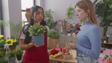Renkli bitkiler ve dekoratif kaplarla çevrili bir çiçekçide sohbet eden iki kadın.