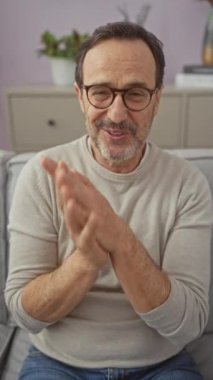 Gülümseyen olgun İspanyol adam evde gözlüklü ve günlük kıyafetlerle oturuyor..